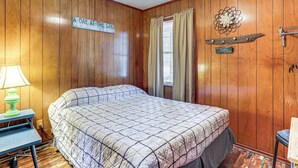 A bedroom with wooden paneling, a bed with a quilted bedspread, a nightstand with a lamp, and a sign on the wall. - Shared Dock & Yard: Greers Ferry Lake Home! (Greers Ferry)