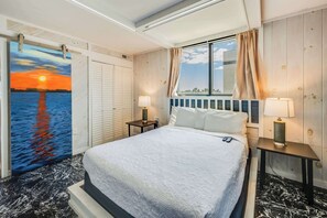 A hotel room with a bed, two bedside tables, a window with curtains, a view of the sunset, and a door with a view of the water. - New Beautifully Remodeled, Meridian Plaza with Pools (Myrtle Beach)