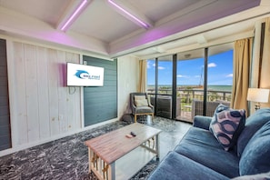 A room with a television displaying a logo, a sofa, a coffee table, and a view of the ocean through large windows.