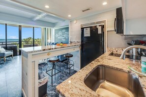 A modern kitchen with a large island, bar stools, a stainless steel sink, and a refrigerator.