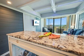 A kitchen with a granite countertop, a TV mounted on the wall, and a view of the ocean through large windows.