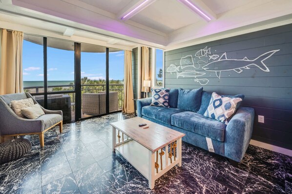 A modern living room with a sofa, armchair, and coffee table, large windows with a view of the ocean, and a wall with a fish mural. - New Beautifully Remodeled, Meridian Plaza with Pools (Myrtle Beach)