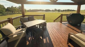 A covered outdoor deck with wooden furniture, a grill, and a view of a grassy field. - Countryside Retreat - Come Relax, Unwind & Cast a Line (LaRue)