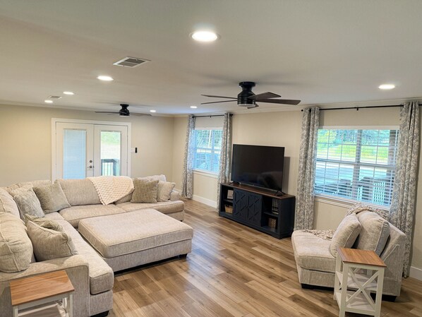 A living room with a sectional sofa, a TV on a stand, a ceiling fan, and large windows with curtains. - Countryside Retreat - Come Relax, Unwind & Cast a Line (LaRue)