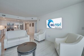 Uma sala de estar moderna com um sofá, poltronas e uma mesa de centro. Uma cozinha é visível ao fundo.