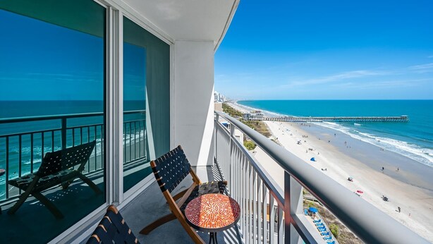 A balcony with a view of a beach and pier, a chair, and a small table.