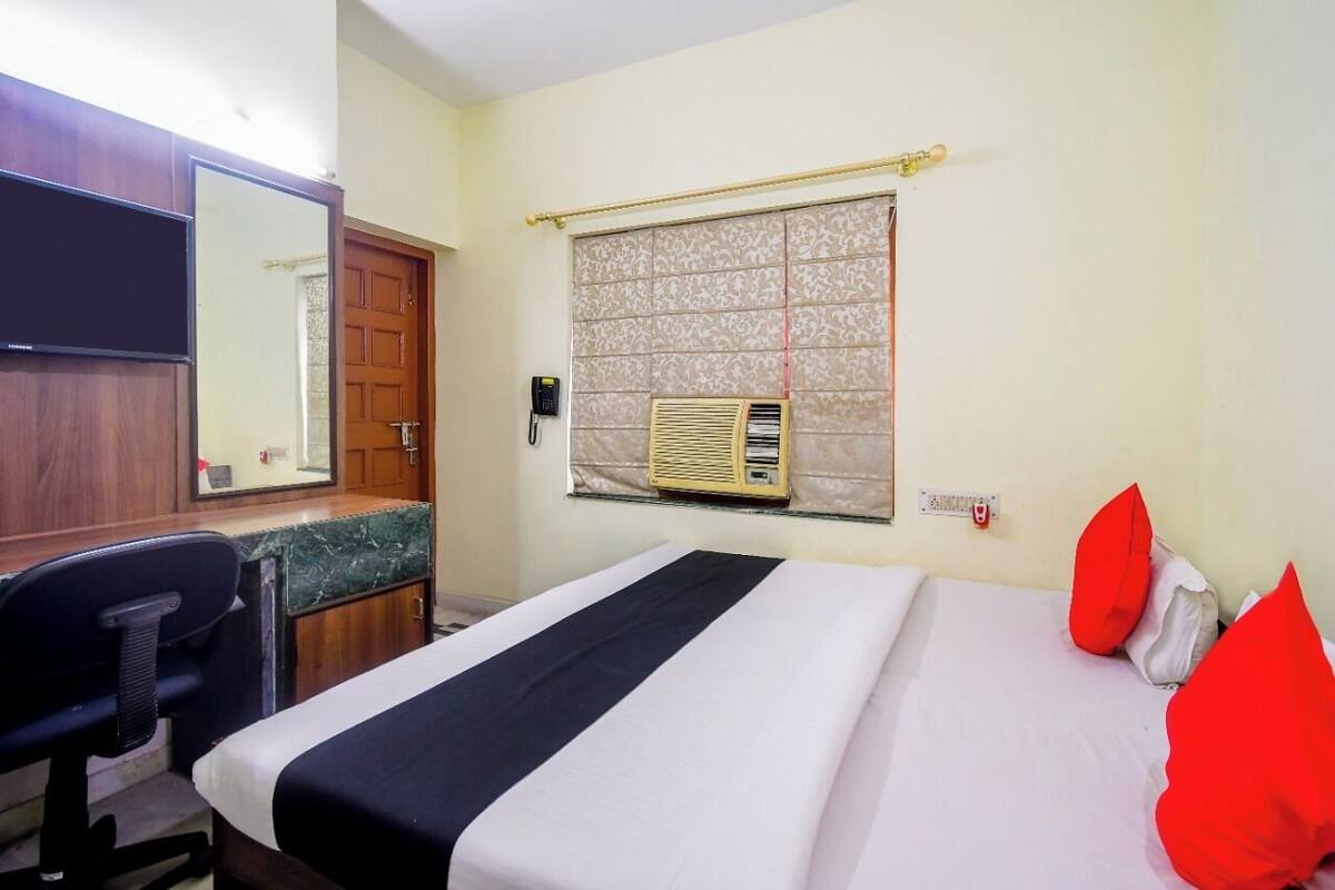 A hotel room with a bed, a desk, a chair, a mirror, a window with an air conditioner, and a telephone.