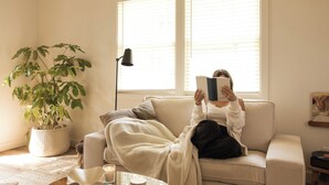 Una persona sentada en un sofá leyendo un libro en una habitación luminosa con un ventanal grande, una planta en maceta y una lámpara.