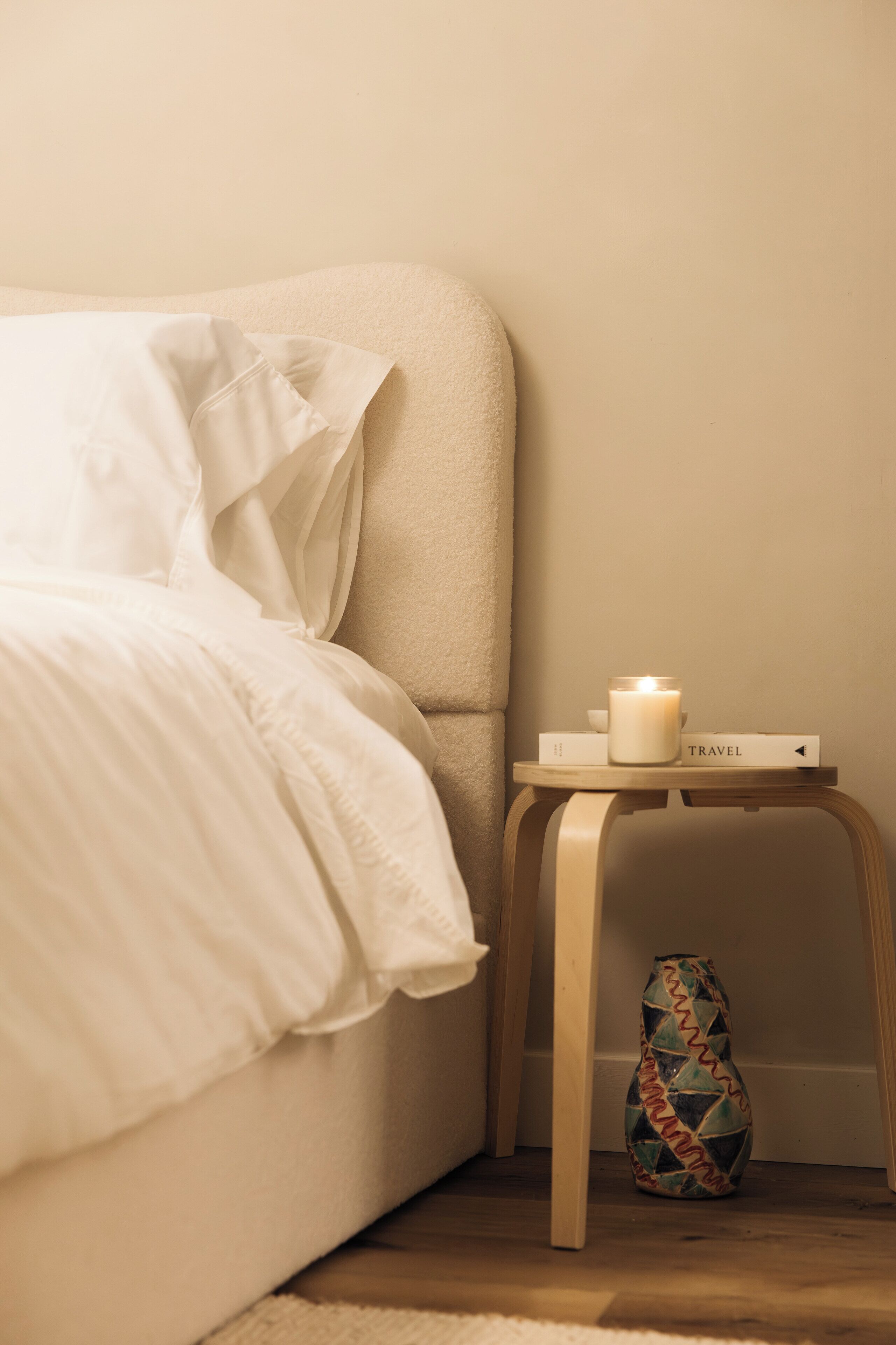 A neatly made bed with white linens, a small bedside table with a candle, a book titled "TRAVEL", and a colorful vase.