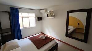 Un lit simple avec une couverture marron et un oreiller blanc, une petite table de chevet, un téléviseur fixé au mur, un miroir et une fenêtre avec des rideaux bleus.
