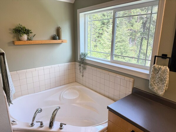 A bathroom with a white bathtub, a window with a view of trees, a wooden shelf with decorative items, and a towel rack with a patterned towel.
