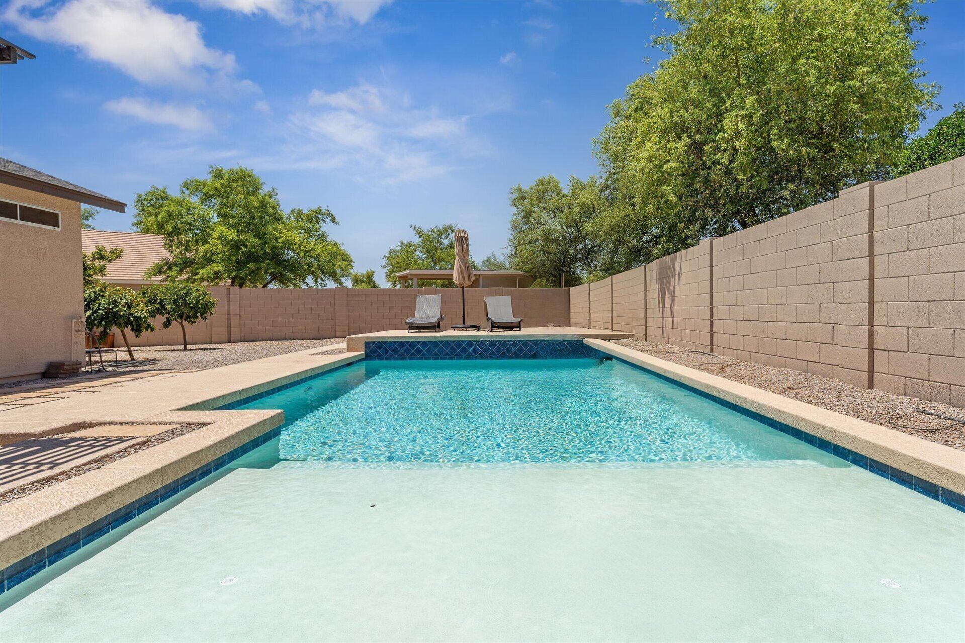 Una piscina rectangular con agua azul cristalina, rodeada por un muro de ladrillo y una terraza de madera. Hay dos sillas reclinables y una sombrilla en la terraza.