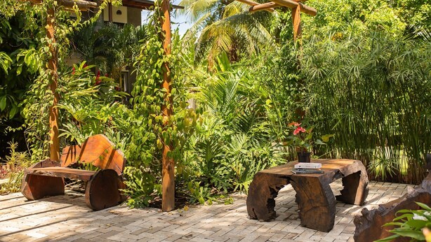 Un patio jardín con muebles de madera rústica, una pérgola y exuberante vegetación tropical.