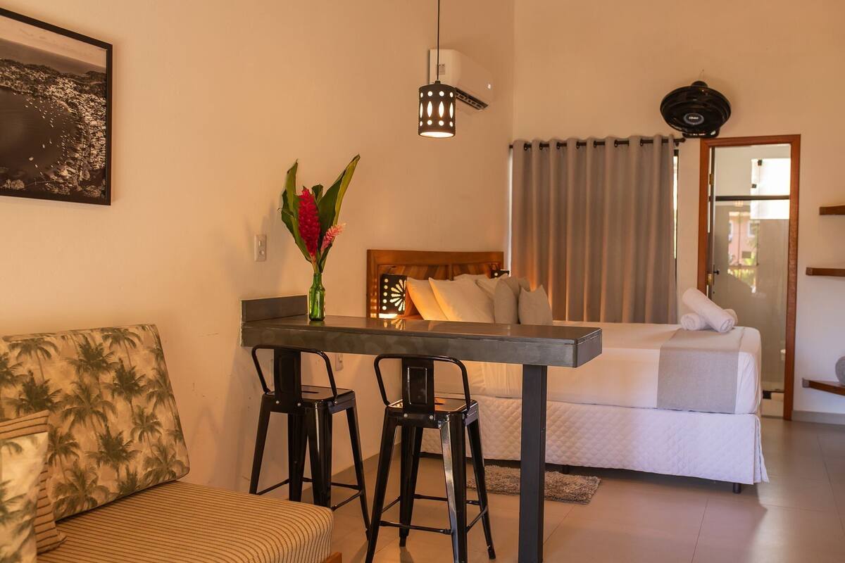 Une chambre à coucher comprenant un lit, une table de chevet, un bureau avec deux chaises, une fenêtre avec des rideaux et un climatiseur fixé au mur.