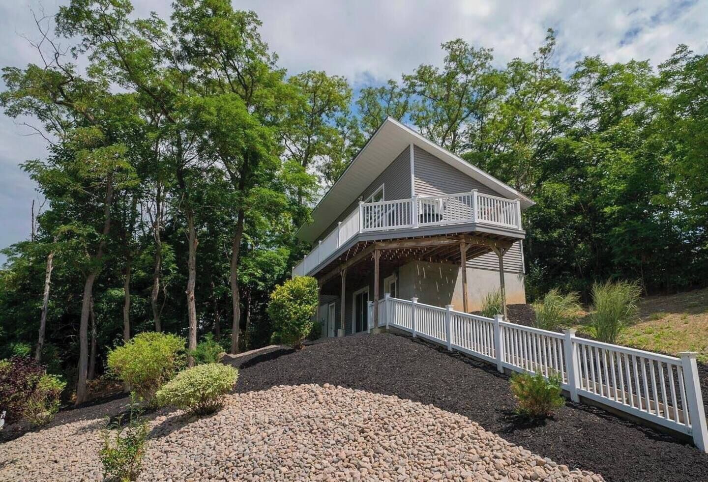 A two-story house with a balcony, surrounded by trees and a gravel driveway.