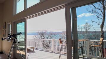 A room with a telescope on a tripod, a balcony with a white railing, and a view of a body of water and mountains.