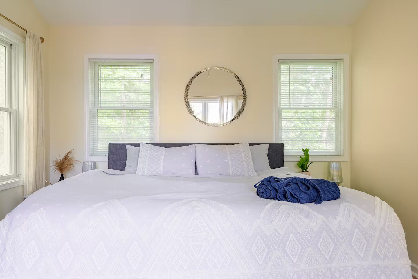 A neatly made bed with white bedding and a blue towel, two windows with blinds, and a round mirror on the wall.
