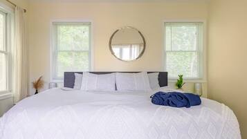 A neatly made bed with white bedding and a blue towel, two windows with blinds, and a round mirror on the wall.