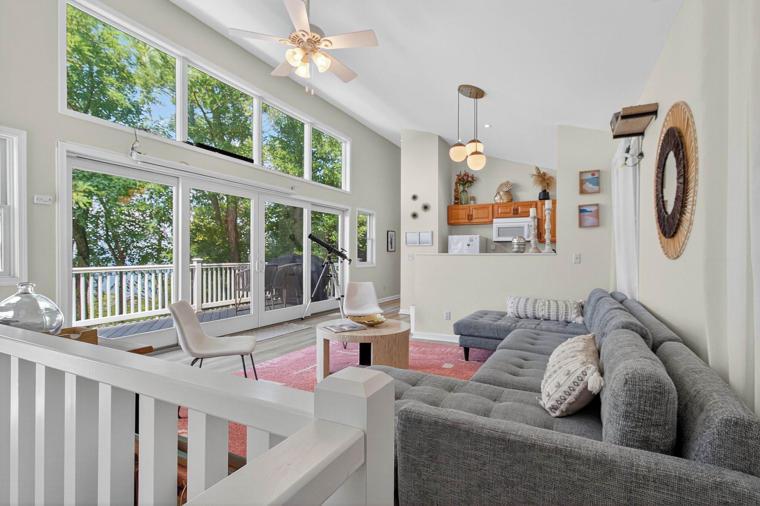 A spacious living room with large windows, a ceiling fan, a gray sofa, and a wooden deck.