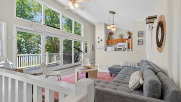 A spacious living room with large windows, a ceiling fan, a gray sofa, and a wooden deck.