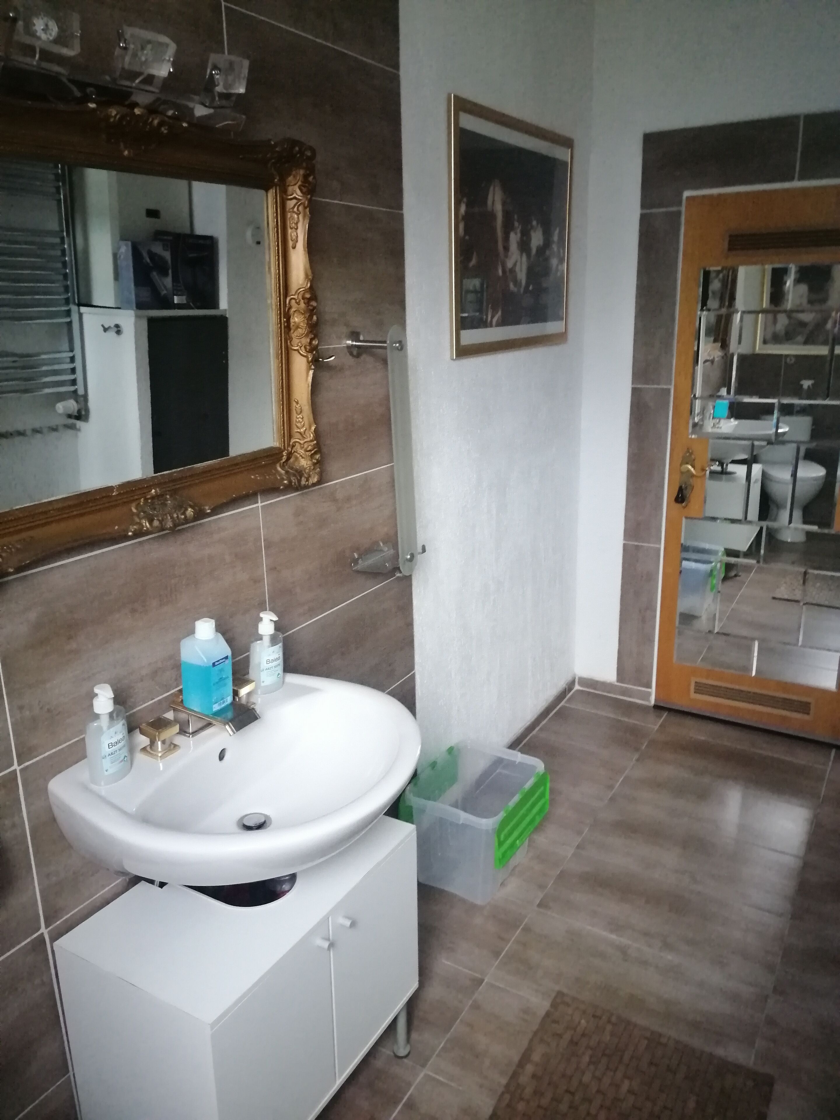 Een badkamer met een witte wastafel, spiegel en betegelde muren. Er staat een groene prullenbak en een wit kastje onder de wastafel.
