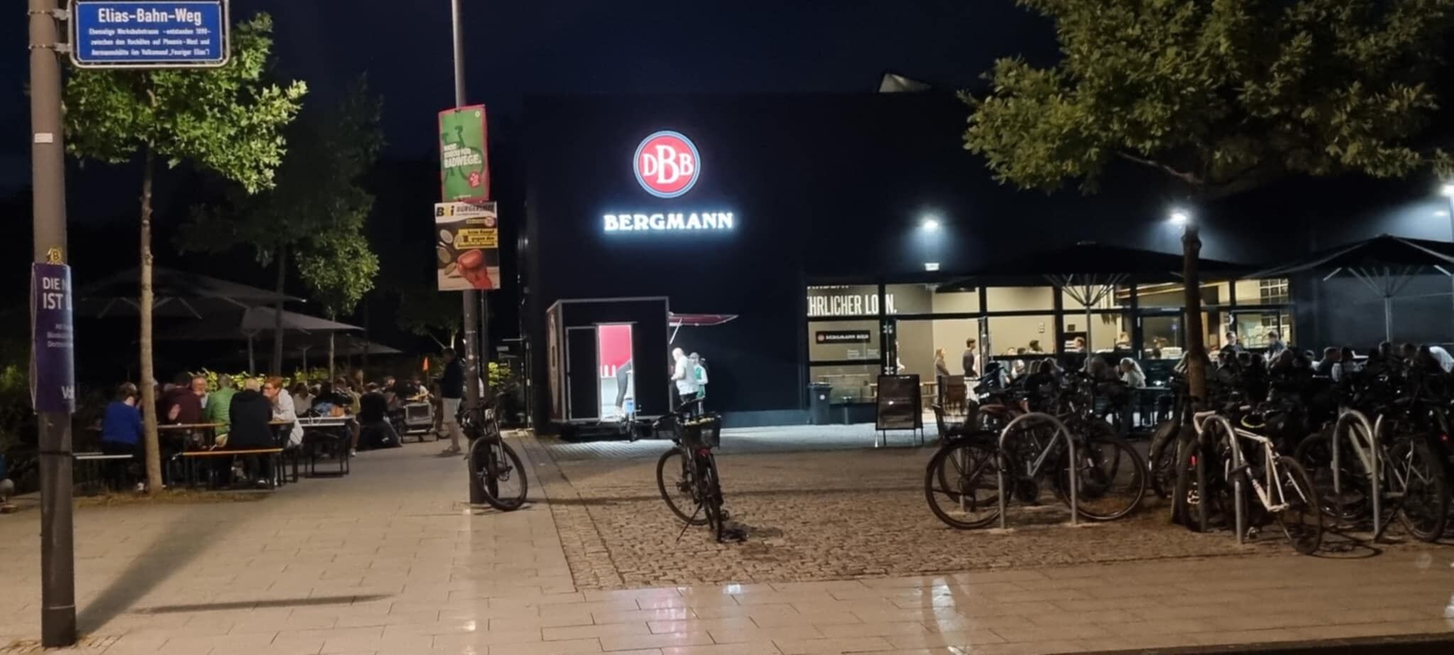 Spazio esterno illuminato di notte, con parcheggio per biciclette e un edificio con un cartello che riporta la scritta "BERGMANN".