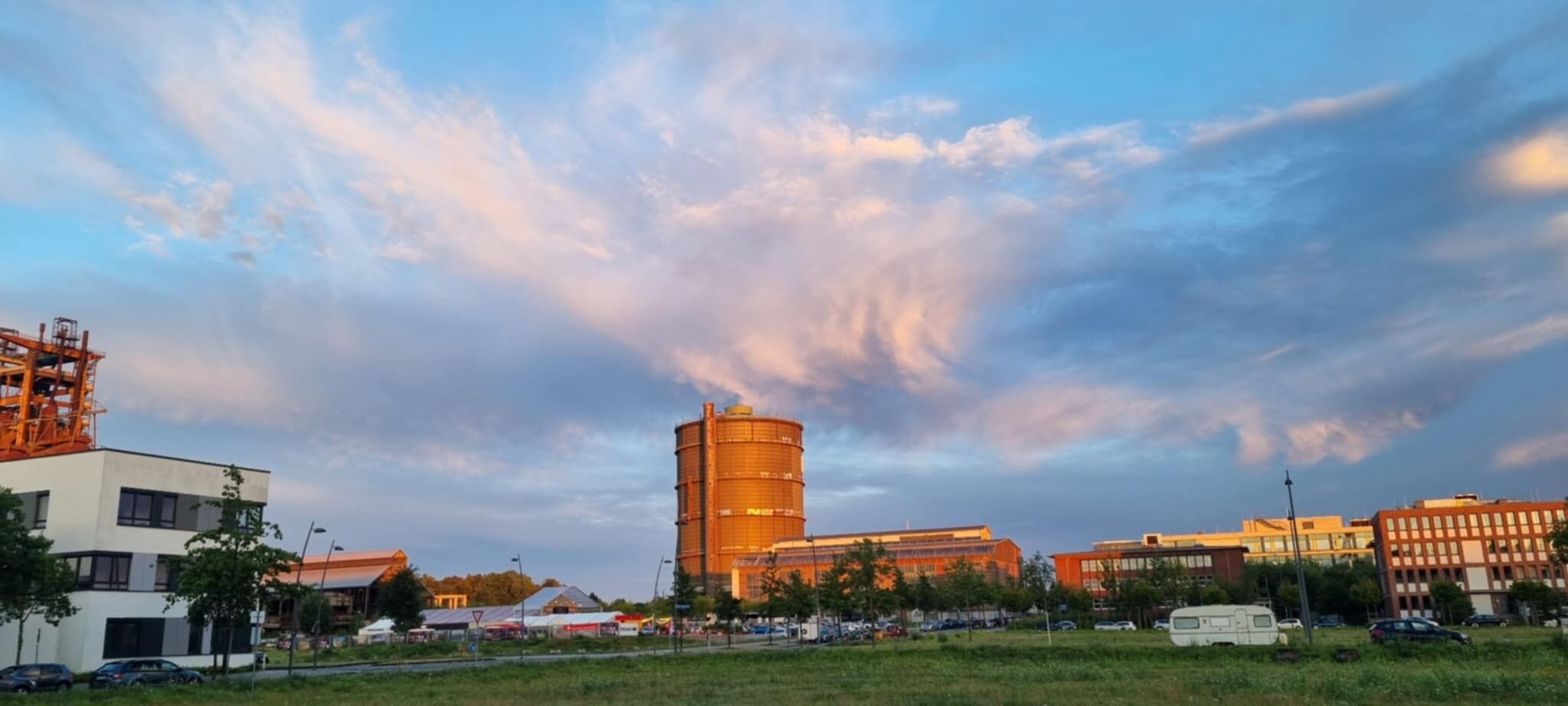 A modern building complex with a prominent cylindrical structure, surrounded by other buildings and a parking area.