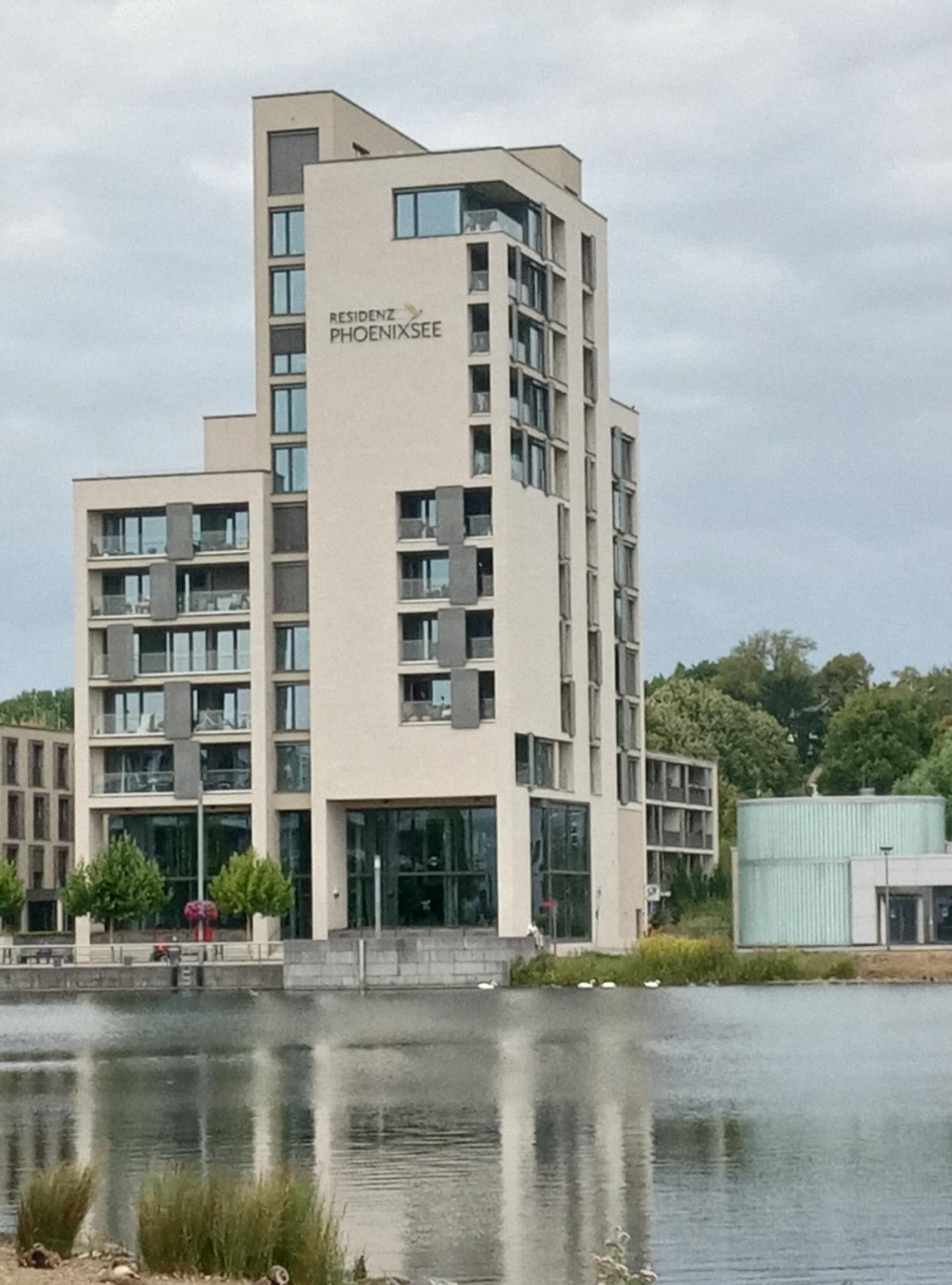 A modern multi-story building with the name "RESIDENZ PHOENIXSEE" on the facade. It features a central entrance with glass doors and multiple balconies.