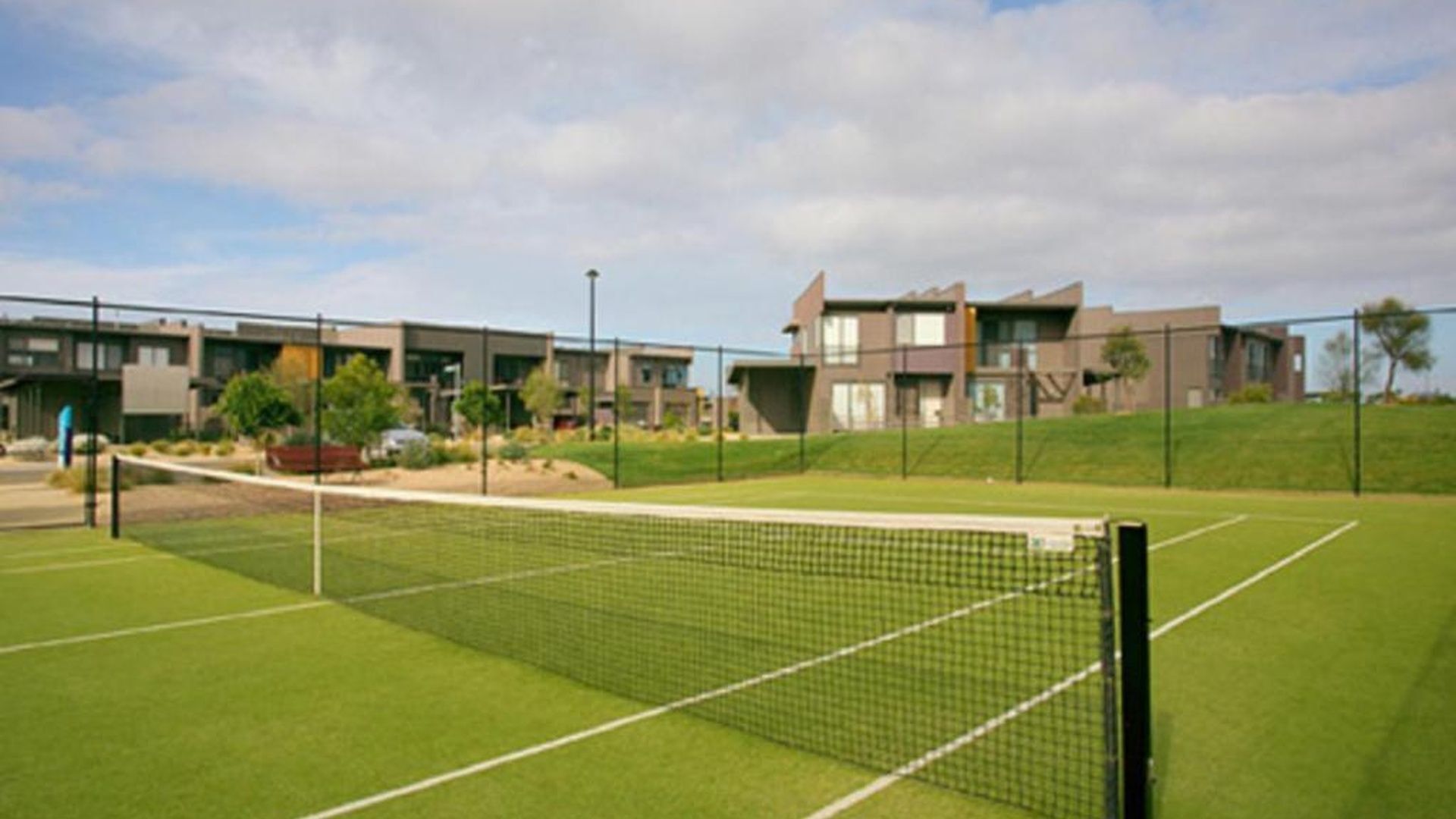A tennis court with a net, surrounded by a fence, and modern buildings in the background.