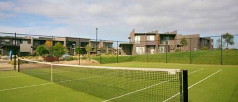 A tennis court with a net, surrounded by a fence, and modern buildings in the background.