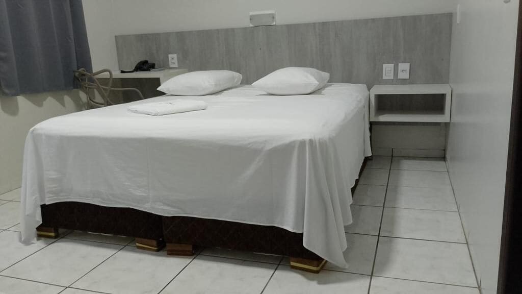 Cama bem arrumada com roupa de cama e almofadas brancas, mesa de cabeceira com abajur e uma pequena estante.