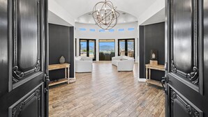 A spacious room with a wooden floor, two white sofas, a chandelier, and large windows offering a view of the mountains.