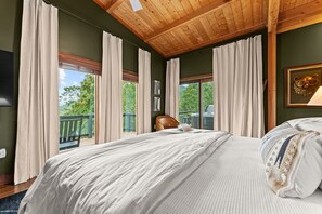 A bedroom with a large bed, wooden ceiling, and a view of a balcony with a railing.