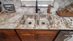 A kitchen with a double stainless steel sink, granite countertop, wooden cabinets, and a window with blinds. - Refuge Hunting Cabin (Elgin)