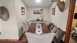 A dining room with a long wooden table, upholstered chairs, and a mounted deer head on the wall. - Refuge Hunting Cabin (Elgin)