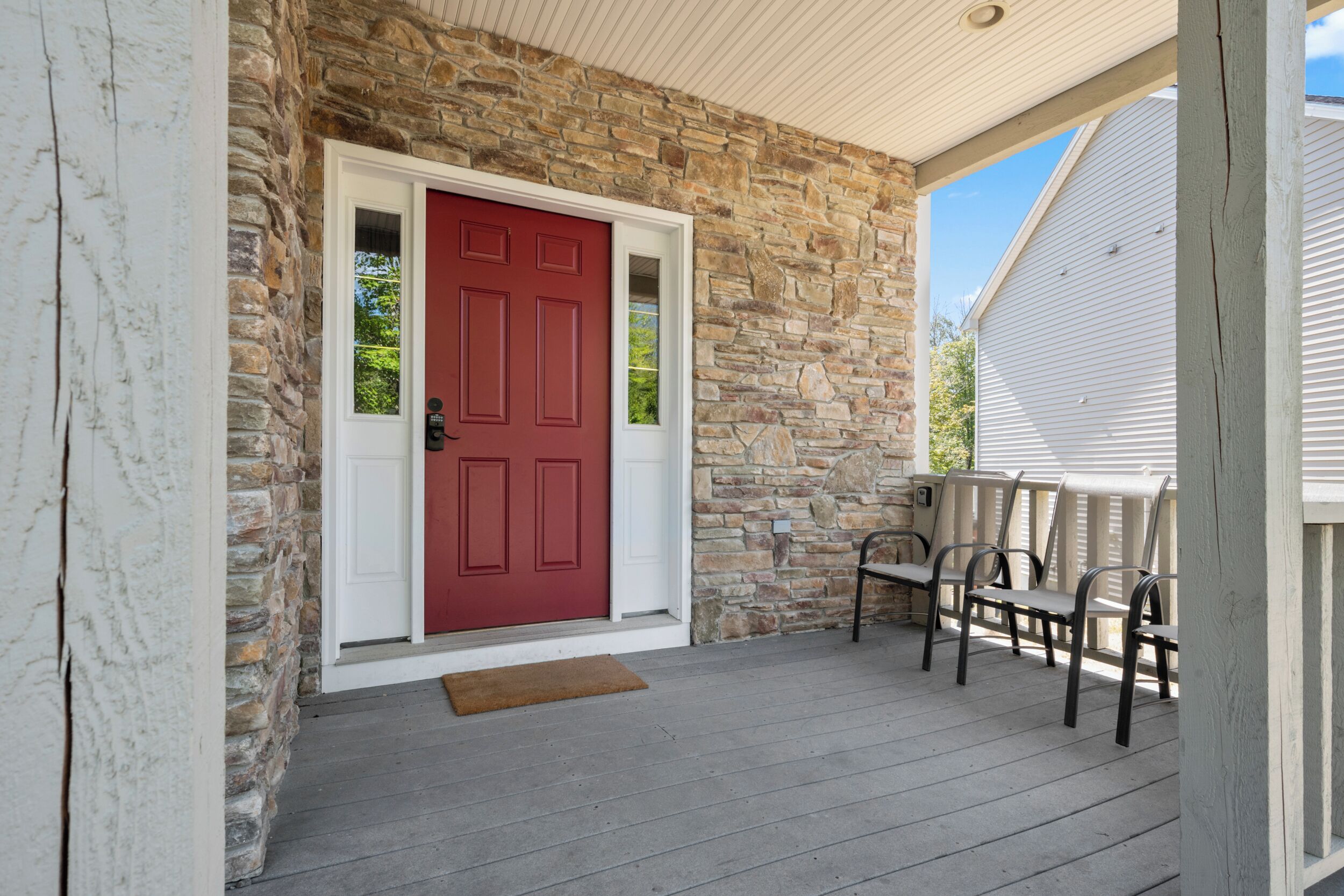 A stone exterior with a red door, a porch with wooden chairs, and a white house in the background.