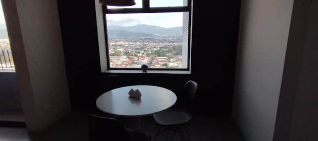 A small room with a round table and two chairs, a window with a view of a cityscape and mountains, and a curtain on one side.
