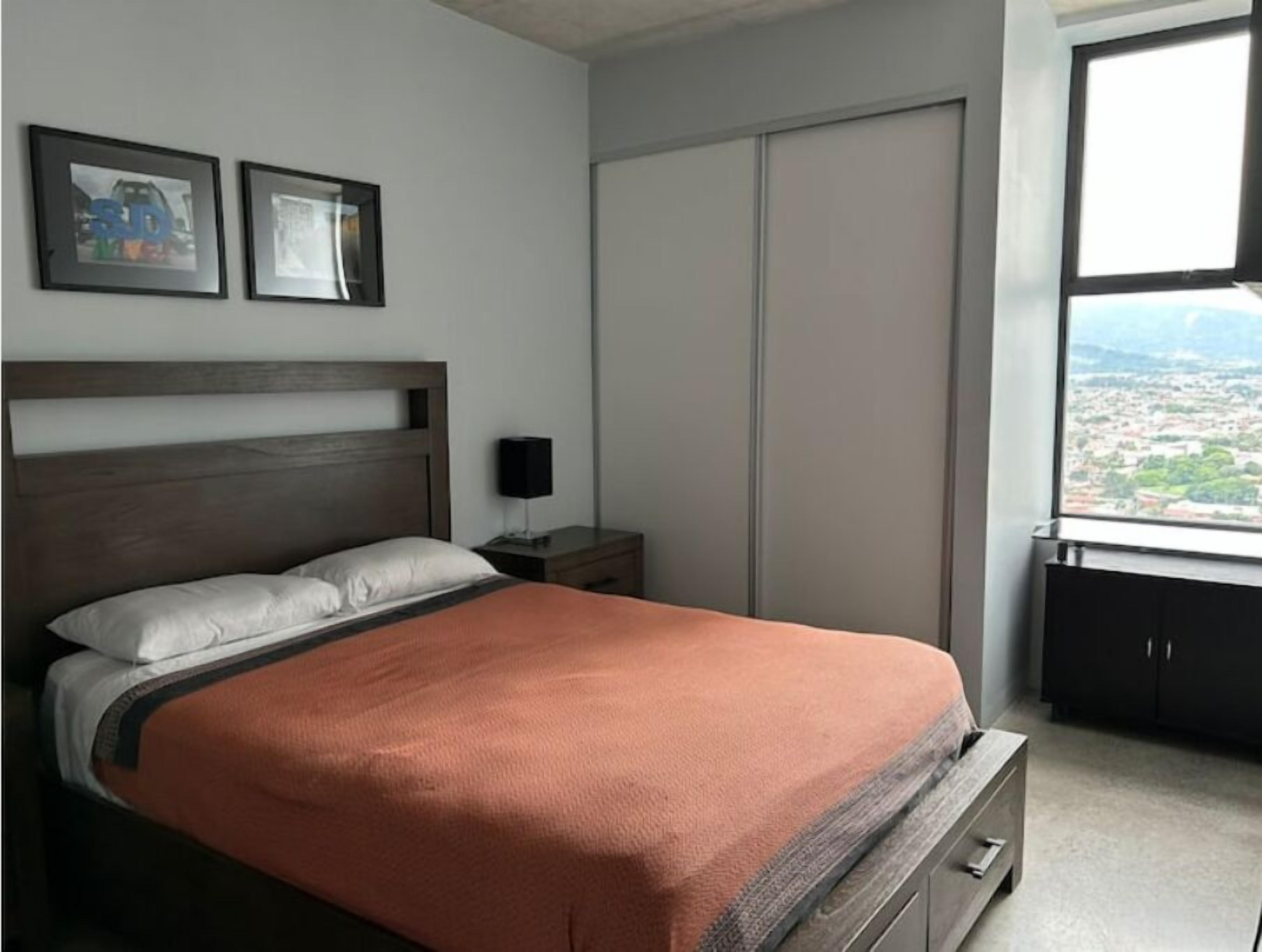 A bedroom with a bed, a nightstand, a wardrobe, and a window with a city view.