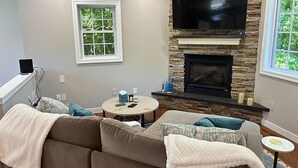 Una sala de estar con chimenea de piedra, televisor de pantalla plana, sofá y dos ventanas.