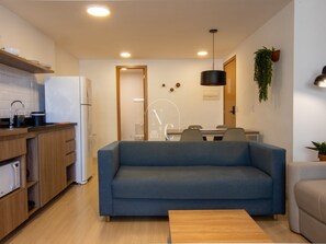 A modern living room with a sofa, a coffee table, and a kitchen area with a refrigerator and cabinets. - Send Co-Living by RNC (Rio de Janeiro)