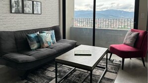 A modern living room with a grey sofa, a red chair, and a coffee table. There are framed pictures on the wall and a large window with a view of mountains.