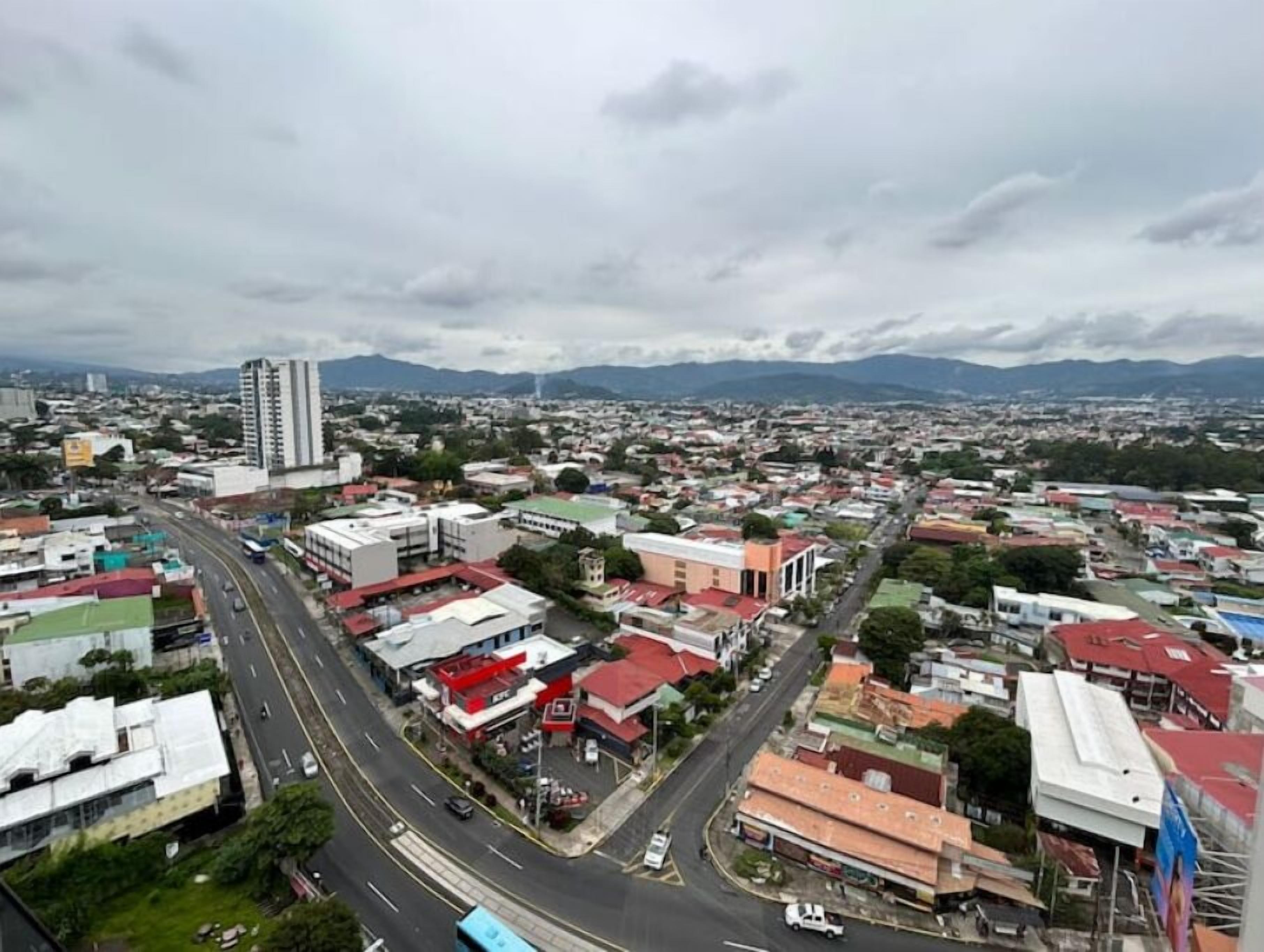 A cityscape with a prominent road, various buildings, and distant mountains under a cloudy sky.