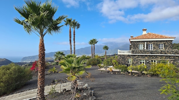 Una casa de piedra con techo de tejas, rodeada por un camino de grava y palmeras, con vistas a un paisaje costero y montañas al fondo.