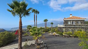 Una casa de piedra con techo de tejas, rodeada por un camino de grava y palmeras, con vistas a un paisaje costero y montañas al fondo.