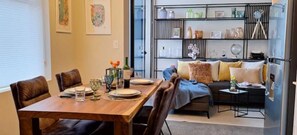 Un coin repas avec une table en bois dressée pour deux personnes, un canapé avec des coussins et une étagère contenant divers objets.