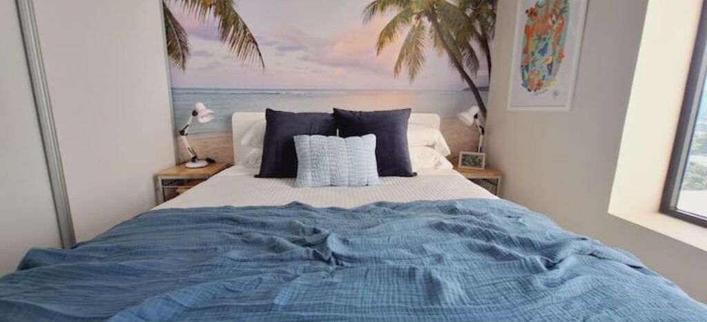 Ein Schlafzimmer mit Bett, Kissen, einem Nachttisch, einer Lampe und einer Wandmalerei mit Strandmotiv.