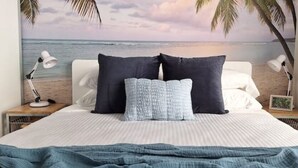 A bed with a white and blue striped bedspread, two dark pillows, and a blue blanket. There are two bedside lamps on wooden nightstands. A beach scene is depicted on the wall behind the bed.
