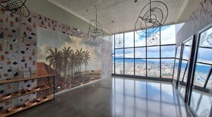 Une pièce spacieuse ornée d’une grande fresque représentant des palmiers et un paysage urbain, de luminaires suspendus modernes et offrant une vue sur la ville grâce à de grandes fenêtres.