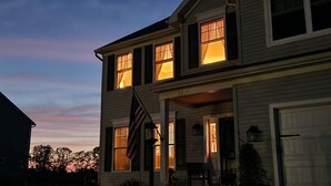A two-story house with multiple lit windows, an American flag, and a porch with chairs. - Mount Nittany View - Your Penn State Home for Every Big Weekend! (Boalsburg)