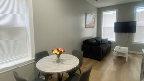 A modern living room with a round dining table, gray chairs, a black sofa, and a flat-screen TV mounted on the wall.